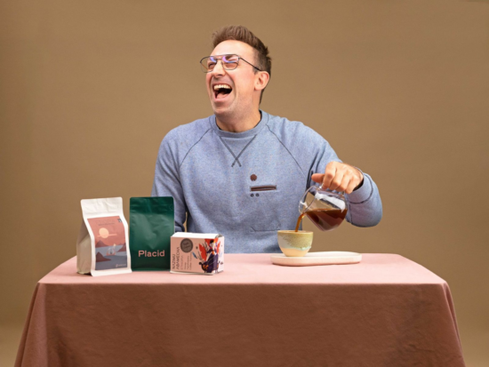 Alex laughs out loud while pouring coffee from a decanter into a pottery cup. He sits at the same pink table and wear glasses and gray sweatshirt with pocket.
