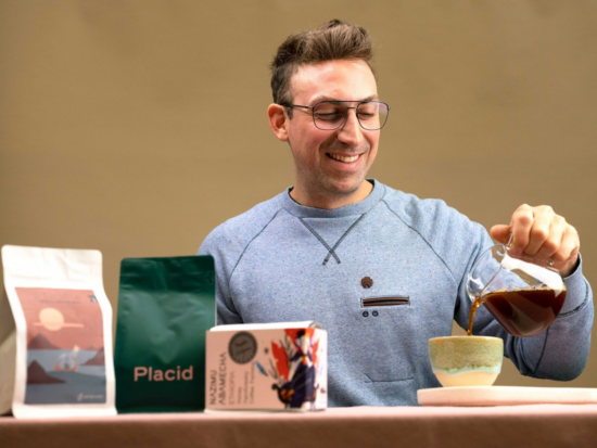 Alex pours coffee into a pottery cup while seated at a table with smooth pink tablecloth. There are three different coffee packages in front of him. Two are bags; one with an illustrated sunset over water, one that simply says PLACID in all caps. There is also a small white box with colorful illustration, a silver seal sticker and letters too blurry to read.