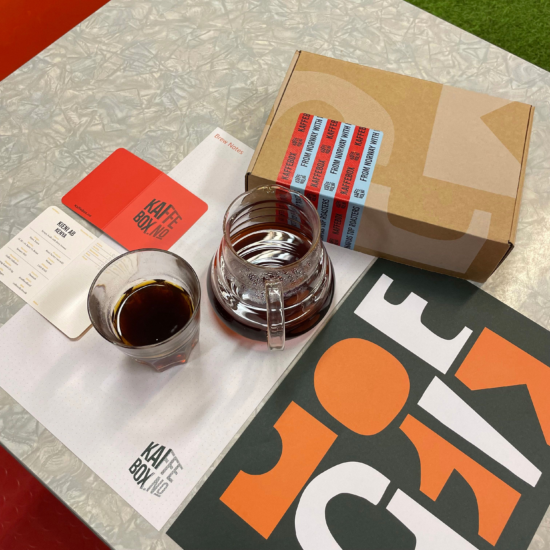 A brown box with sticker label for Kaffebox. Before it is a glass decanter with brewed coffee and a double rocks glass with some coffee inside. There is a little red booklet with KAFFE BOX.NO printed on the front. There is also an abstract black, white and orange print on the table beside.