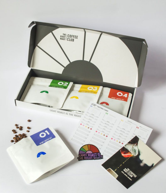 The same long white box with lid, with four small white flat coffee bags inside. Each is labeled with a different number and color (1 is blue, etc.) There is a little rainbow sticker that says "light roast is the right roast." There is also a coffee club booklet with an image of coffee being poured into a white cup with a glass decanter. They have included a taste sheet describing the coffees in the box.