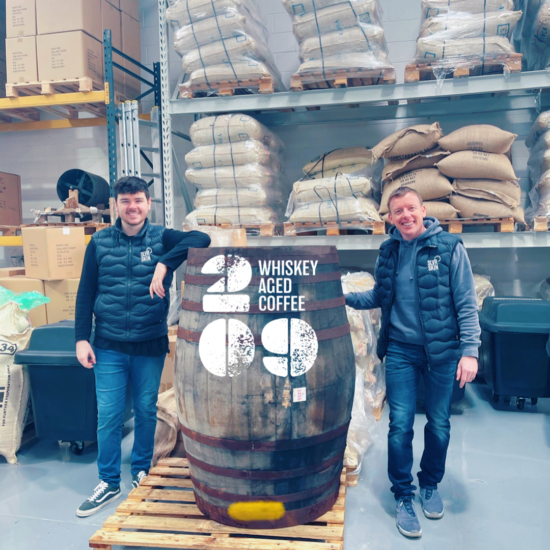 Two men stand in a warehouse next to a huge barrel labeled whiskey aged coffee. Behind them, on pallets and a metal industrial shelf, are stacked huge bags of green coffee beans.