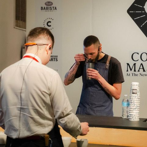 Davide is tasting something with a spoon out of a metal cup. Tattoos are visible on his forearms. In front of him someone is tasting something from a ceramic cupping vessel.