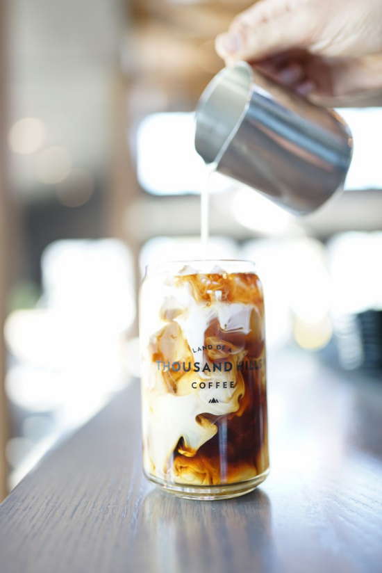 A close up of a metal pitcher pouring cream into a can glass of cold brew coffee that reads Land of a Thousand Hills.