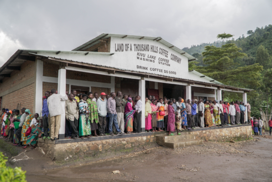 A large gathering of people on the porch and around a building. It is the outside of the Land of a Thousand Hills Kivu Lake washing station. The sign on the building labels the place and says Drink Coffee, Do Good.
