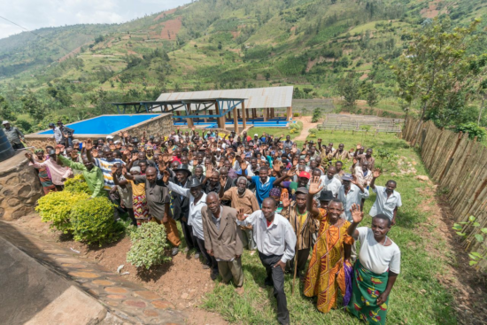 A large group photo of the Ruli Mountain coffee farmers of Rwanda. There are dozens of men and women, both young and old; some are in traditional Rwanda dress and others in suits and Western attire. Many wear hats. They are raising their right hands in greeting and smiling. Behind the group, which is outside on the grass, is a small mountain and a covered porch.
