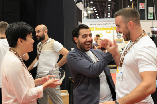 LAF representatives (two men with lanyards, one in a suit jacket) grasp hands in victory after receiving an award at World of Coffee in Milan. A woman holds out a circular clear glass award, about six inches around, towards the victors.