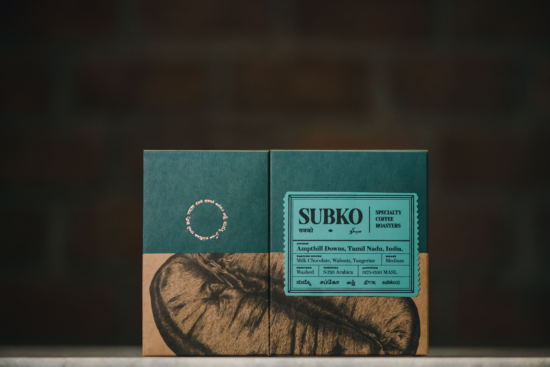 Two Subko coffee boxes fit together to show the entire illustration of a close-up coffee bean. The drawing is hyper-detailed, with half on the front and half on the side of the box, so that it wraps around the left side. There is a big blue product stamp, designed to look like old train tickets from India.