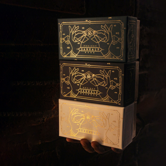 Стопка из трех коробок Sankalp размером с чайную коробку, на которых изображены руки двух женщин, выполненные сусальным золотом.  В каждой руке по большому кофейному зерну.  На руках есть браслеты, а также граница и фон из минимальных геометрических фигур, таких как бриллианты.