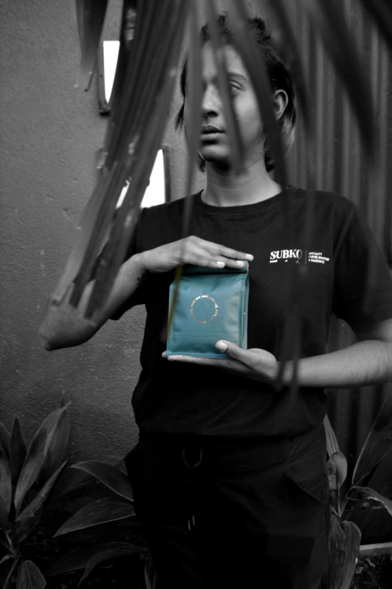 Модель із напівзачепленим волоссям позує з сумкою Subko перед собою, одна рука зверху та друга внизу.  Перед її обличчям пальмове листя.  Вона одягнена в чорні штани та футболку з логотипом Subko.