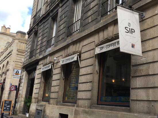 The White flag sign of Sip, hanging from the side of a grey concrete building with tall windows painted with coffee signs for espresso and pastries.