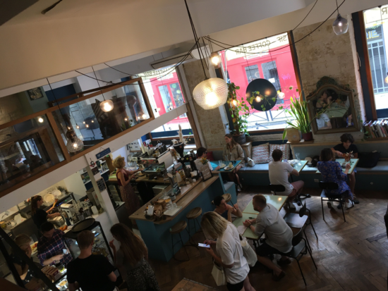A view from the second story of Sip Coffee Bar, where you can see the blue counters with wood tops, Art on the brick walls and zig zag pattern wood floors below. On the left the clear glass partition wall of the second floor is visible, with wooden railing.