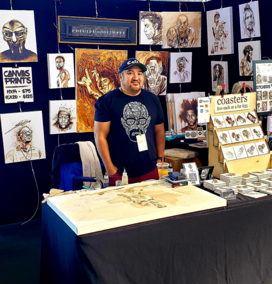 Jarold poses smiling, with hands in pockets in his art stall. He wearsa ball cap and a Breaking Bad t shirt with Walter White's head. Laid out on the table is a big canvas he is working on, and a display of painted coasters. On the wall behind hang many different portraits of celebrities drawn in coffee, such as Prince, Biggie, Bruce Lee and Michael Jordan.