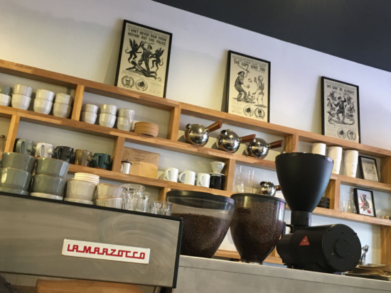 Behind the espresso machine at Black List, a white wall features built in minimalist wooden shelving with bowls, mugs, and sinister old fashioned ink drawings of devils and skeletons.