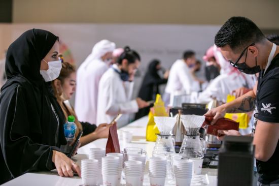A woman in a headscarf watched a man in glasses weigh out coffee for a pour over demonstration. Many people can be seen in the background watching baristas work. The table is stacked with white paper cups and coffee bags in different colors.