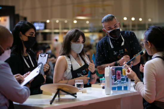 A woman in glasses demonstrates something in a plastic case with different colored liquids or solubles. Judges wearing masks and carrying clip boards listen and ask questions. 
