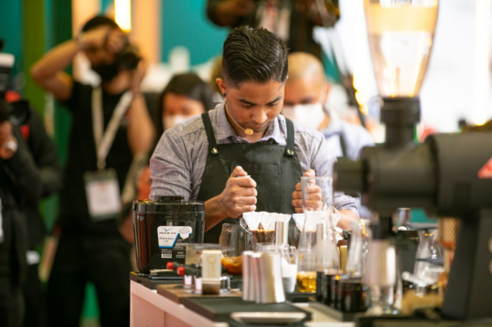 A barista with a headset, wearing a smooth canvas apron, holds two whisks in place while working on a coffee presentation. An assortment of scales and containers and other coffee paraphernalia block the view of his drink in process.