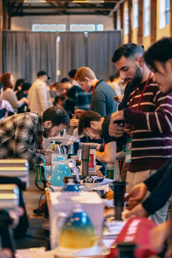 A long table has baristas crammed around it, all men, working on their coffee recipes, They have gold and blue kettles, AeroPresses, cupping vessels and spoons all across the table. They each wear a lanyard.