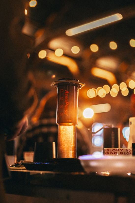 Stylized close up of an empty AeroPress on a table. There is a mug near it that says "No Glory." There is also a Comandante grinder behind it. Lights twinkle and blur in the background.