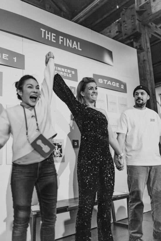 An announcer in a one-shoulder sparkly jumpsuit holds the hands of Jibbi, which is raised, and the hand of the runner up,  who wears a beanie. 