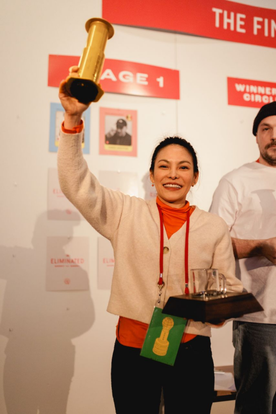 Jibbi Little, on stage, holds up in her her right hand the gold championship trophy, which is shaped like and AeroPress. She wears an orange turtleneck with a buttoned pink cardigan on top, and wears he hair pulled back. 