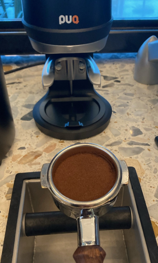 A portafilter with tamped espresso rests on a knock box in front of a Puqpress.