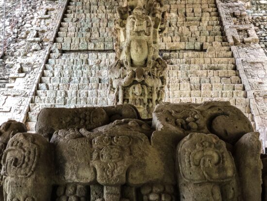 A well-worn statue of a god or priest at the Mayan ruins. It appears to be holding a vessel in its hands. It wears a large stone headdress and stands atop a pedestal with swirling designs that look like snakes and fruits.