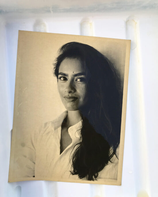 Una foto de retrato en tonos sepia de una mujer joven con cejas oscuras y cabello largo sonriendo directamente a la cámara. Lleva una camisa blanca con botones.