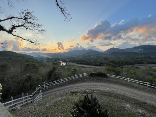 A cloudy sunset on a hill. The hill is surrounded by trees, and other higher hills can be seen in the distance. A gravel driveway is directly before the camera, and a white picket fence runs behind it.