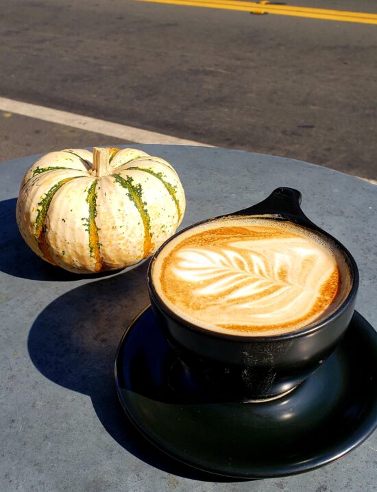 A black ceramic mug with saucer contains a latte, complete with latte art, and a white pumpkin with specks of green and orange stripes sits on the concrete surface behind it.