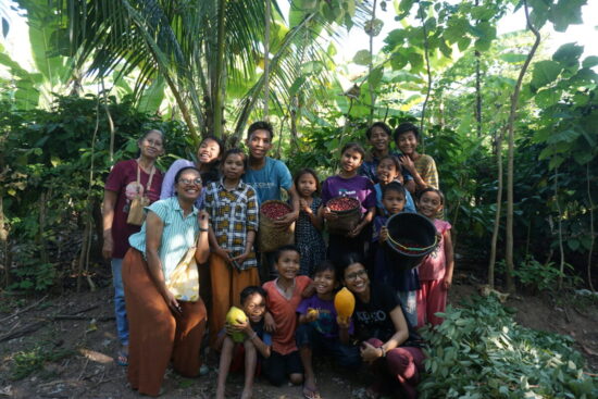 Mahniwati and a group of 13 kids and 2 other adults gather for a group photo. Some hold up large fruits, while some carry baskets with coffee cherries. Behind them is vibrant green tropical trees and foliage.