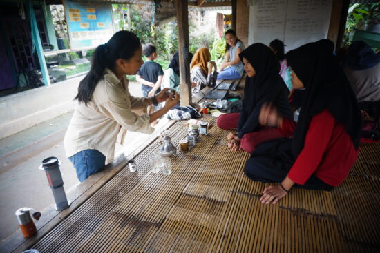 A woman in jeans and a button up shirt trains some younger women on coffee beans. The woman stands while the younger women sit on mats, wearing head scarves. 