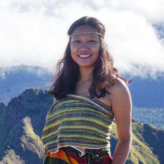 A photo of Mahniwati of Kon Bayan Coffee beaming in front of a mountainous landscape. She wears colorful stripes in a wrap top and skirt, and has a thin headband holding back her long black hair.