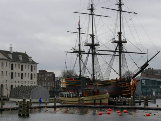 A replica of a Dutch ship in the harbor at Amsterdam. It has a high prow, masts, gold and brown painting on the side. metal reinforcements, and windows in the back cabin.