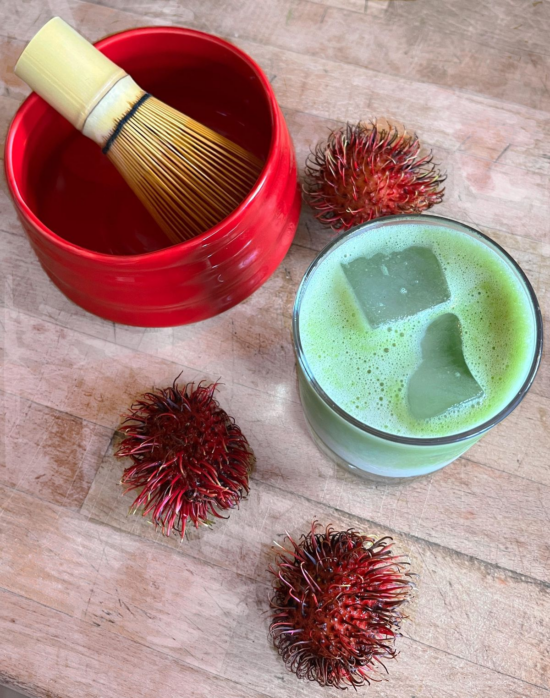 An image of a red matcha bowl with a bamboo matcha whisk inside it. There is a glass of iced matcha beside the bowl. Scattered on the table are three small rambutan fruits, still whole. 