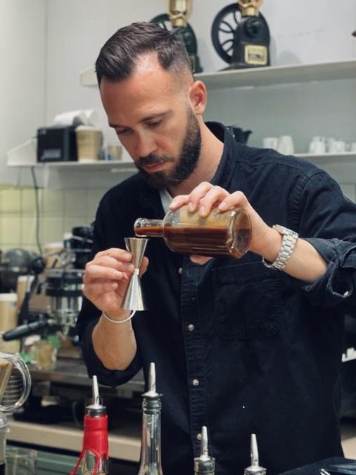Christos pours cold coffee from a glass bottle into a jigger to measure it for a cocktail. Behind him is an espresso machine. In front of him is a selection of syrups for bar use, all with pour spouts attached.