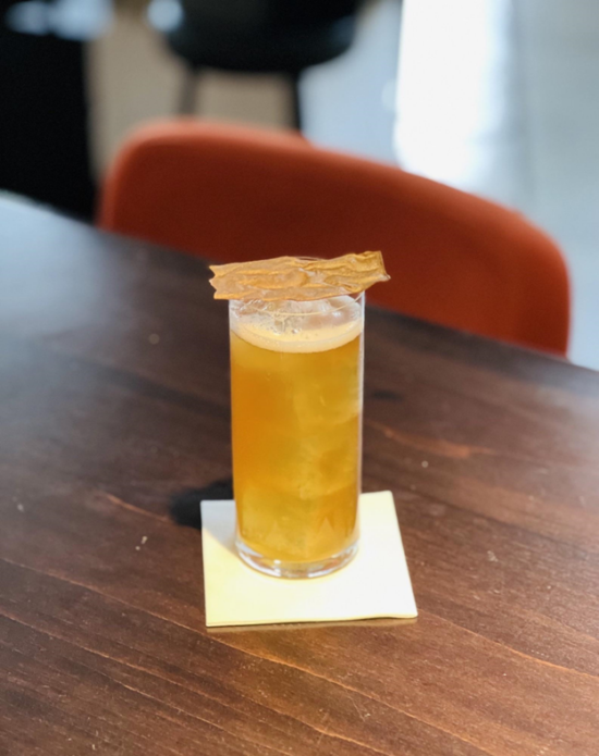 In a Collins glass, a sparkling black zero-waste honey coffee cocktail has an amber color and a papery sugar garnish on top.