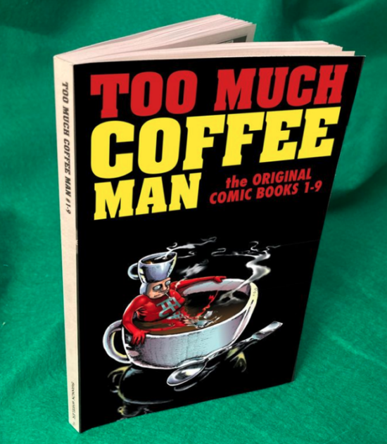 A copy of the new TMCM one-volume paperback book, with all caps title in yellow and red and black cover. On the cover is TMC Man lying in a giant cup of coffee, similar to a hot tub, with a huge spoon lying beside it. He is gripping the sides and smoking a cigarette.
