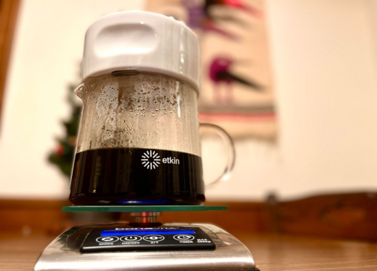 The Dripper on top of a scale with coffee brewing through it. The etkin symbol (similar to a sun, identical to the raised lines of the inside of the dripper) and the world etkin is printed on the side of the clear glass carafe.