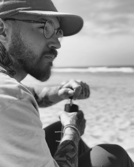 Dávid Stefanik, also known as Bayreesta, in a black and white photo. He is grinding beans with a handheld grinder at the beach. He wears a corduroy ball cap, has tattoos, glasses, and a short beard. 