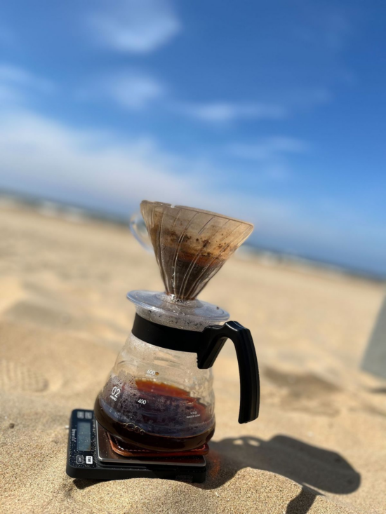 Dávid Stefanik's Hario V60 brewing on the beach. The device, on a glass carafe, sits on top of a coffee scale set on the sand. The background of sandy beach and blue sky is blurry.