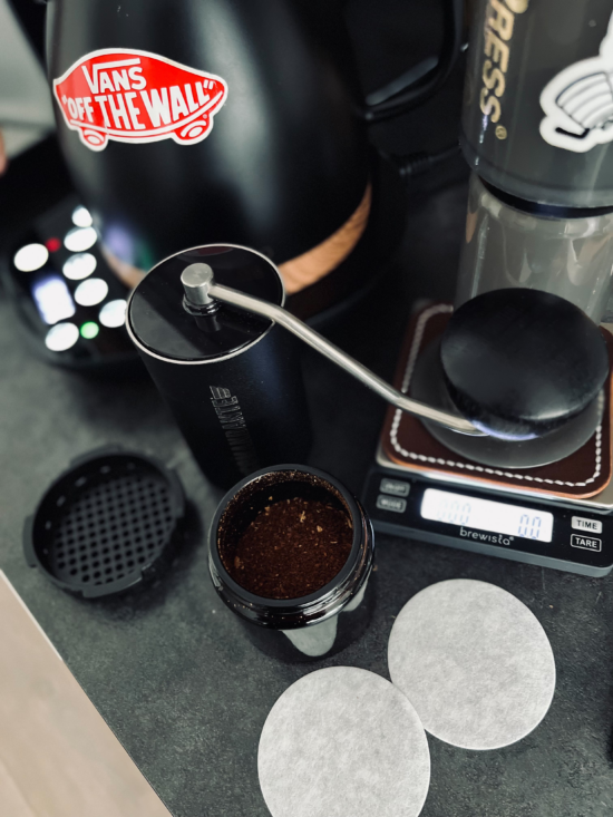 Dávid Stefanik's travel kit includes coffee grounds inside a Comandante, two AeroPress filters. a scale, AeroPress, and a black electric kettle all sitting on a gray counter.