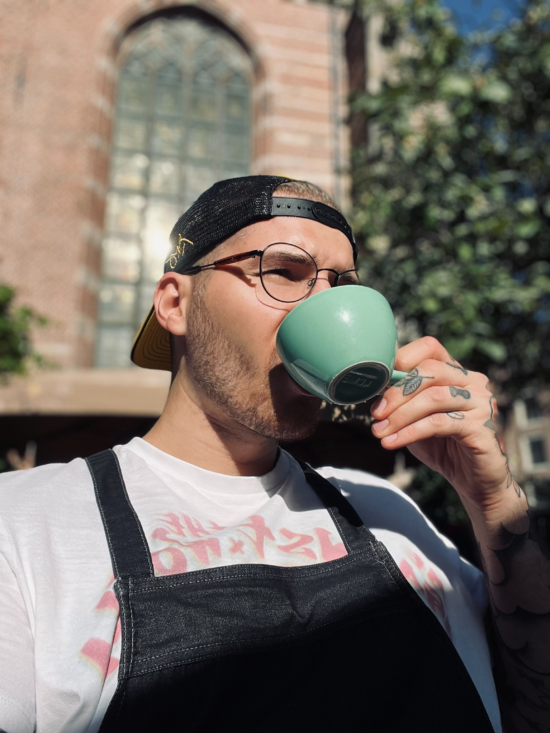 Dávid sips from a light blue ceramic cup, outside a brick building. He wears a denim apron and a backwards baseball cap. 