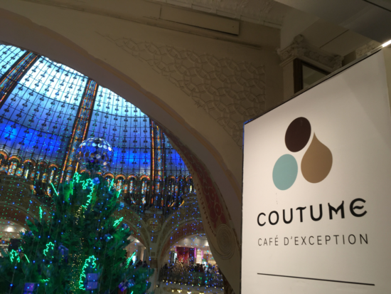 The interior of the shopping mall housing Coutume cafe. The enormous glass dome ceiling is blue. You can see arches above large pillars and a big Christmas tree with blue and green lights.