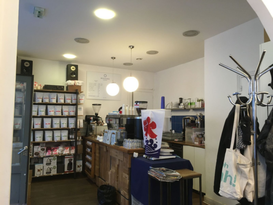 Inside Hexagone, a shelf for retail coffee, a reclaimed wood counter, coffee brewing equipment, and chocolate bars are illuminated by big circle lights.