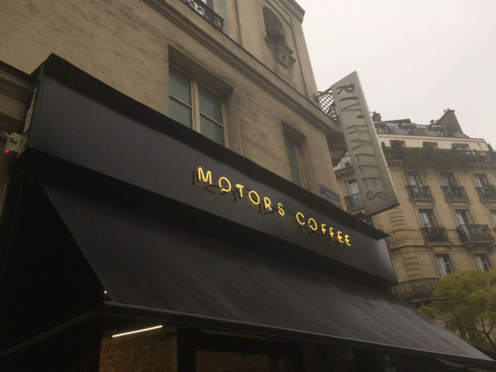 The outside of Motors Coffee in Paris. The sky is a gloomy gray. The awning above the shop is black, with the shop logo protruding in yellow neon. Behind the building are tall 19th century apartment buildings.