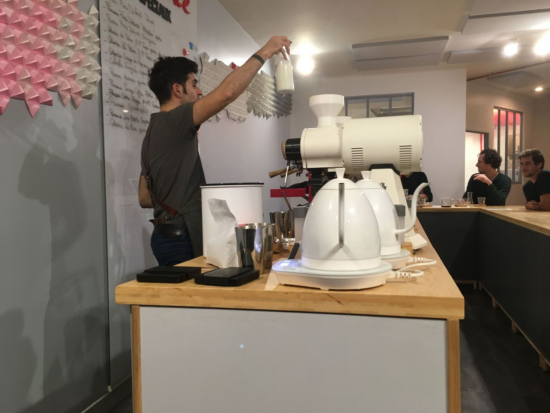 Joachim behind the coffee counter holds up a white pitcher above his head while he talks to his guests. They sit around him at a U shape bar.