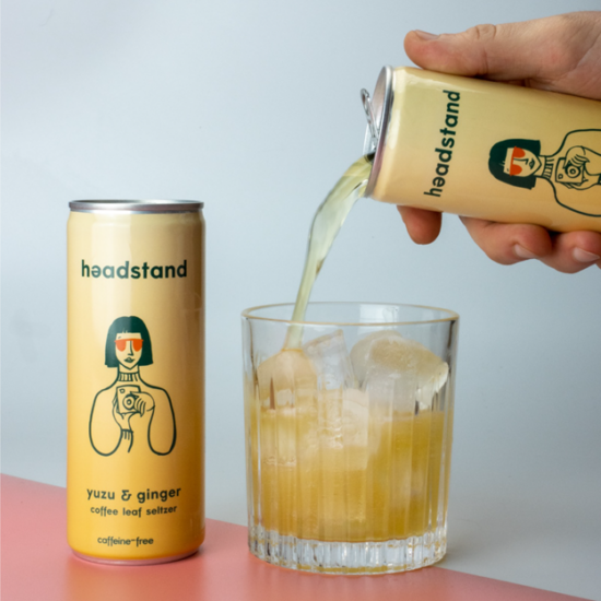 A can of yuzu and ginger coffee leaf seltzer from headstand. It's yellow with a stylized drawing of a person holding a film camera, with a bob haircut and orange sunglasses. A can of the seltzer is also being poured into a rocks glass and is light orange in color.
