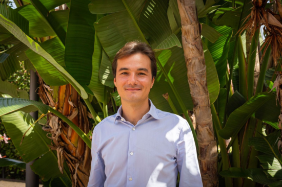 Портрет Андреа, стоящей перед гигантскими тропическими листьями.  Он носит рубашку на пуговицах и улыбается. 