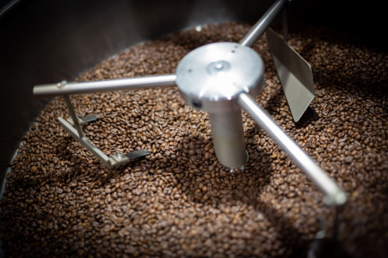 Pogled unutar pržionice kave, sa zrncima koja su već srednje pržena, i tri ruke od nehrđajućeg čelika na kotaču koji okreću zrna.