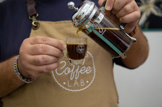 A barista in a tan colored canvas apron pours French press coffee into a glass espresso cup. 